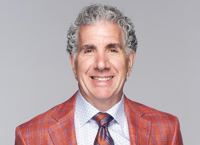 Craig Button has curly grey hair and is wearing an orange tartan blazer over a dress shirt and tie.