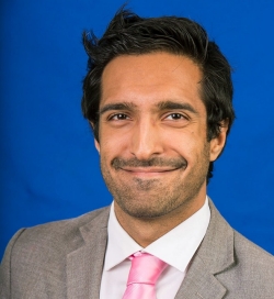 Salim Valji is a sports journalist posing in front of a blue background. He has dark hair and is wearing a blazer over a tie and collared shirt.