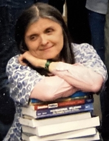 Susan sits with a pile of books on her lap. She leans on her arms that are crossed on top of the book pile and smiles slihtly while looking to the left.