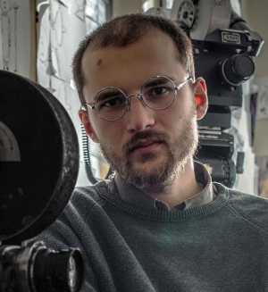 Alex is wearing a collared shirt under a sweater and round wire-rimmed glasses. He has brown hair in a buzz cut. He poses next to film cameras.