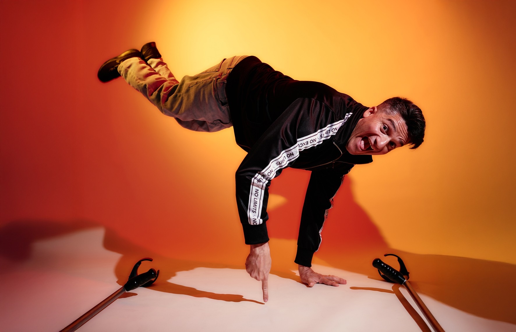 Break dancer Luca Patuelli poses by standing on one hand and one finger. He smiles with his mouth open in front of an orange-hued backdrop.