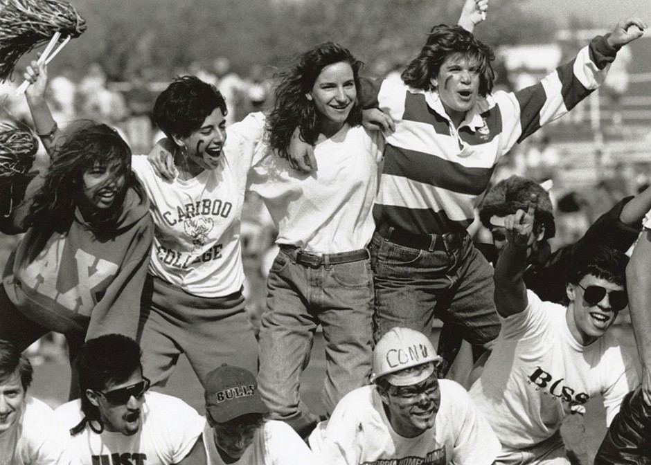 Four students stand on the back of four other students at a football game. they are wearing jeans, sweatshirt and have their faces painted with team colours.