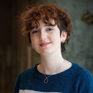 Ruby has short curly red hair and wears a blue and grey sweater.