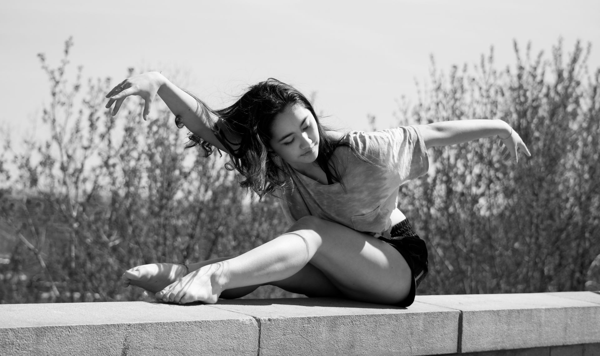 Emma is balanced on the top of a brick wall in a graceful ballet pose with her arms extended one in in front, one behind.