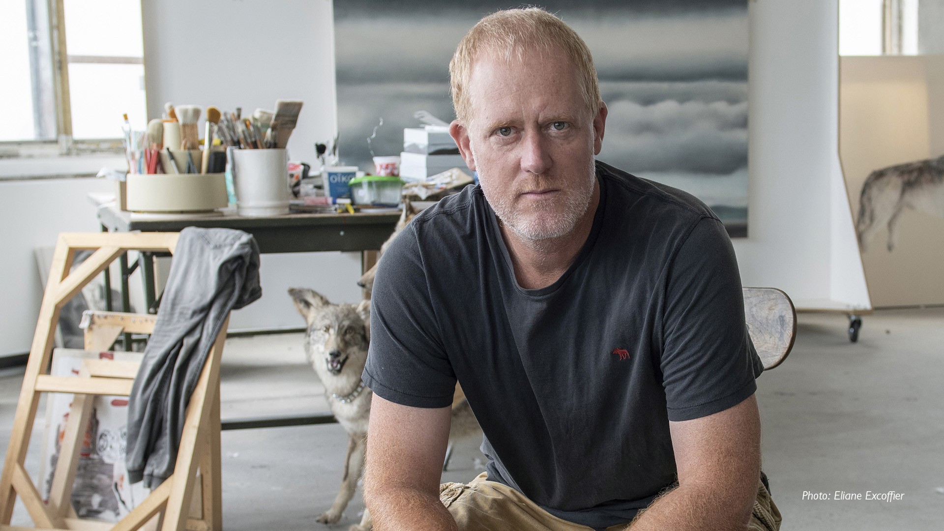 Marc Séguin, BFA 95 Marc is sitting in his painting studio. He has close-cropped red hair and is wearing a black T-shirt.