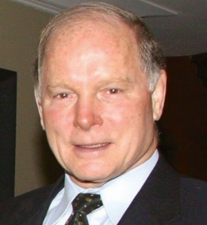 He has short, light hair and eyes that appear brown in the photo. He wears a suit.
