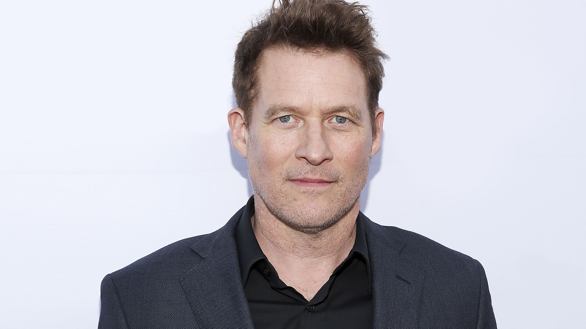 James has short brown hair swept up in the front. He is wearing a black shirt under a dark grey blazer.