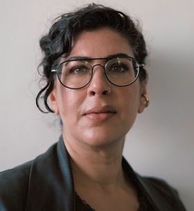 Swapnaa has her dark hair pulled back with one long tendril falling over her face. She wears black metail-rimmed glasses and a black suit jacket.