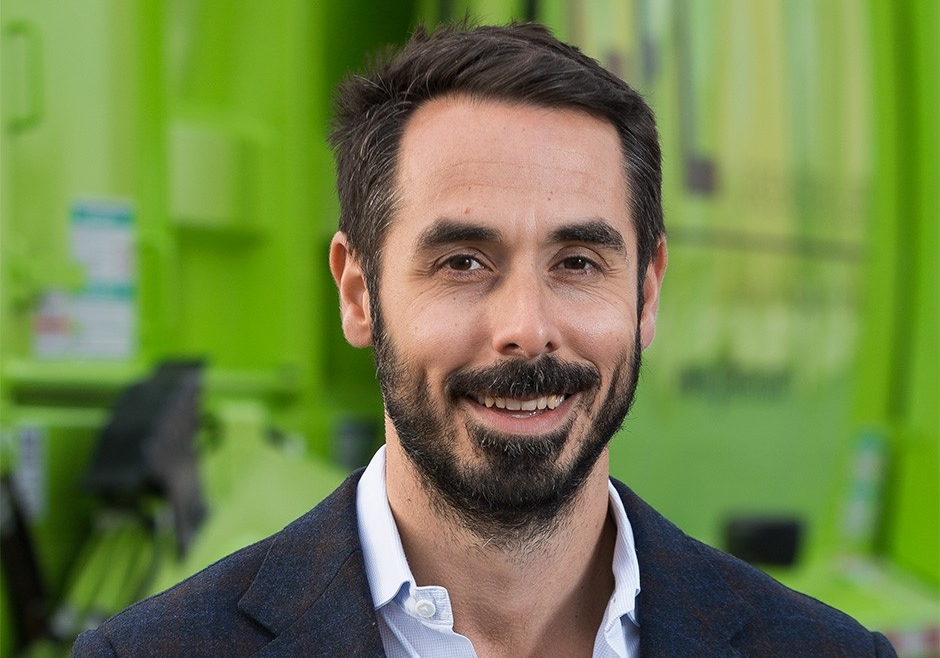 Luke Pelosi Against a bright green background, Luke has short dark hair and a trim dark beard. He wears a dark suit jacket and white shirt.
