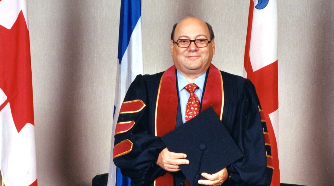 Francesco Bellini is wearing a convocation robe and is standing in front of three flags.