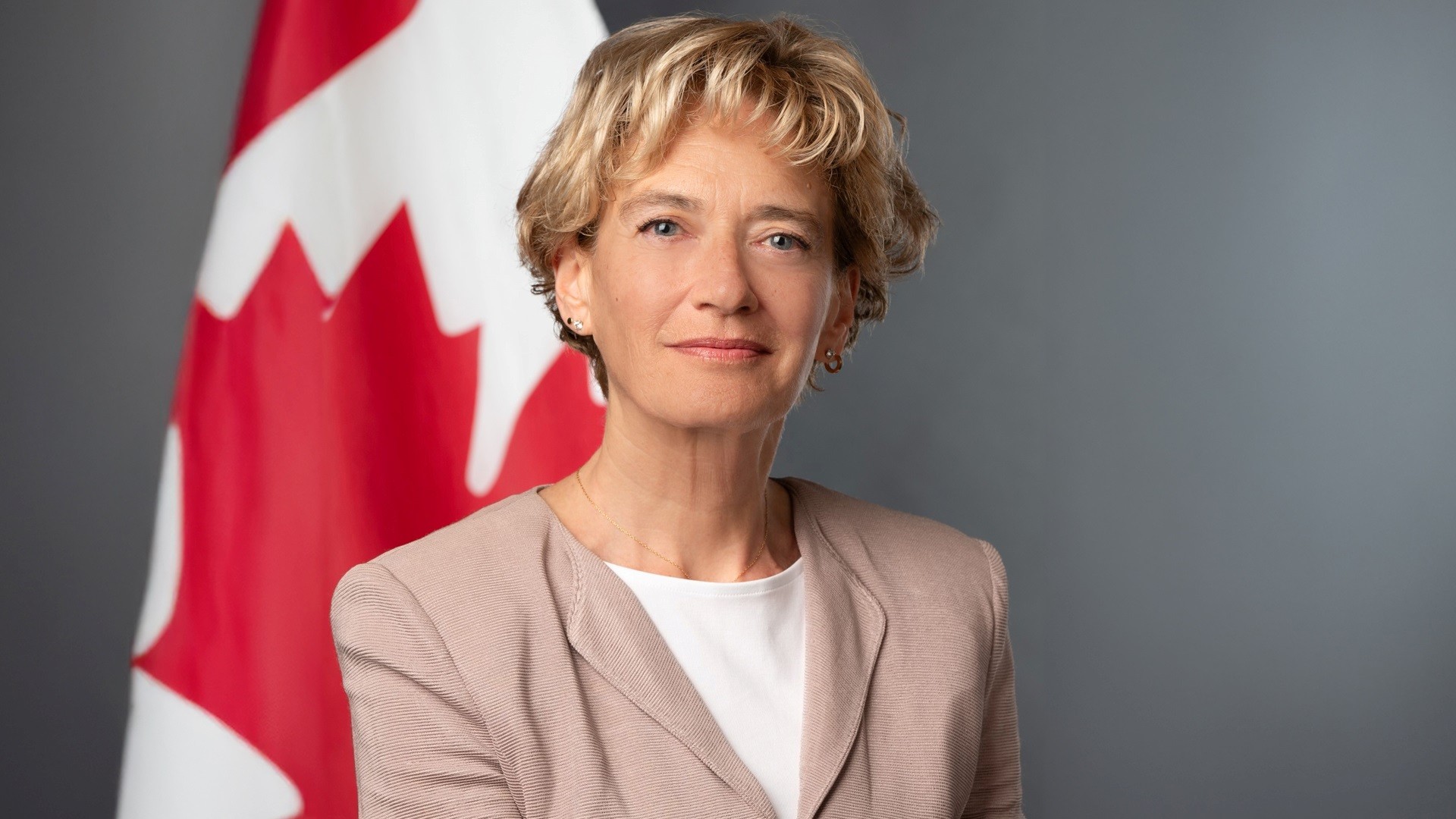 Joyce Napier stands in front of a Canada flag and is wearing a rose coloured blazer over a white shirt.