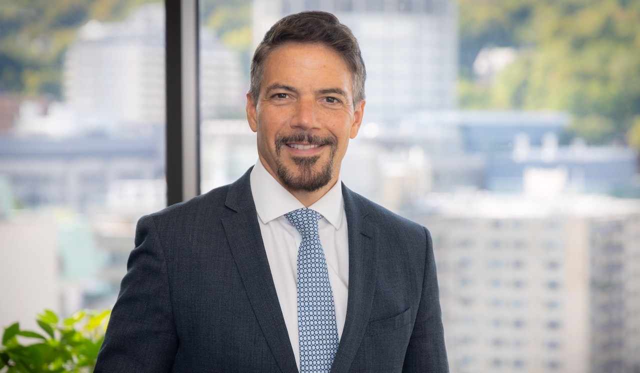A man with short dark hair and beard is wearing a suit and is standing in front of a large window with buildings in the background..