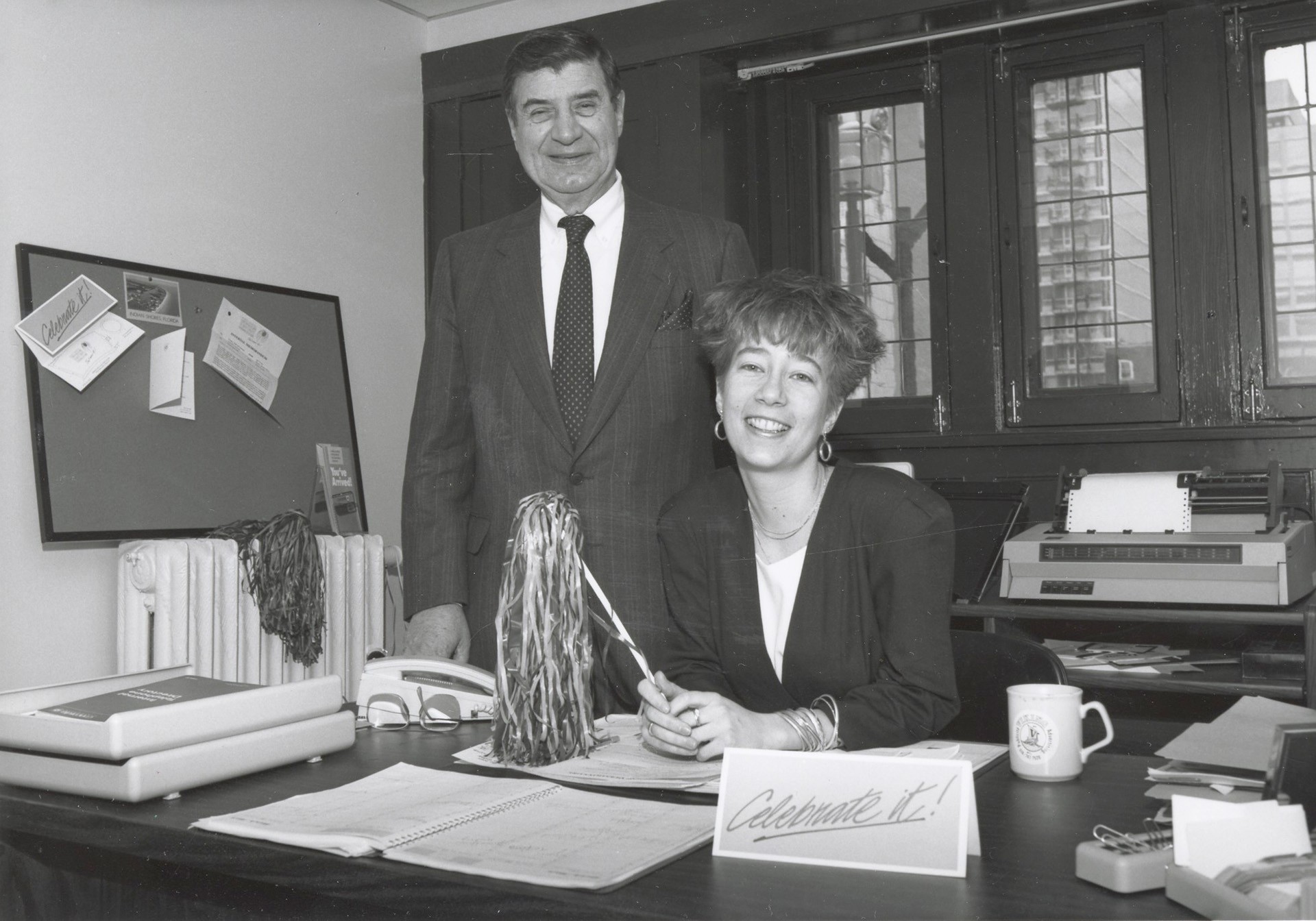 Leisha sits at ther desk and volunteer John Economides stands next to her.