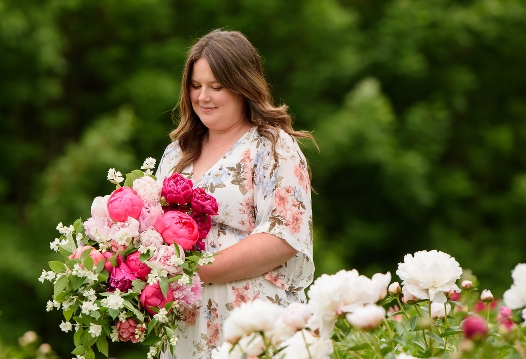 A woman with long, brown hair is wearing a floral dress and is holding a bouquet of pink and white peonies, surrounded by a field of peonies.