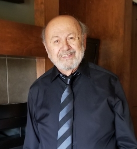 A man with a white beard is smiling and wearing a dark dress shirt with a blue striped tie.