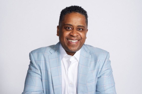 Gary is wearing a light blue suit and a white shirt. He is seated at a white table and smiles.
