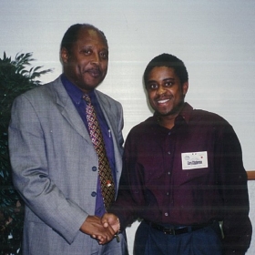Two men smile at the camera while they shake hands.
