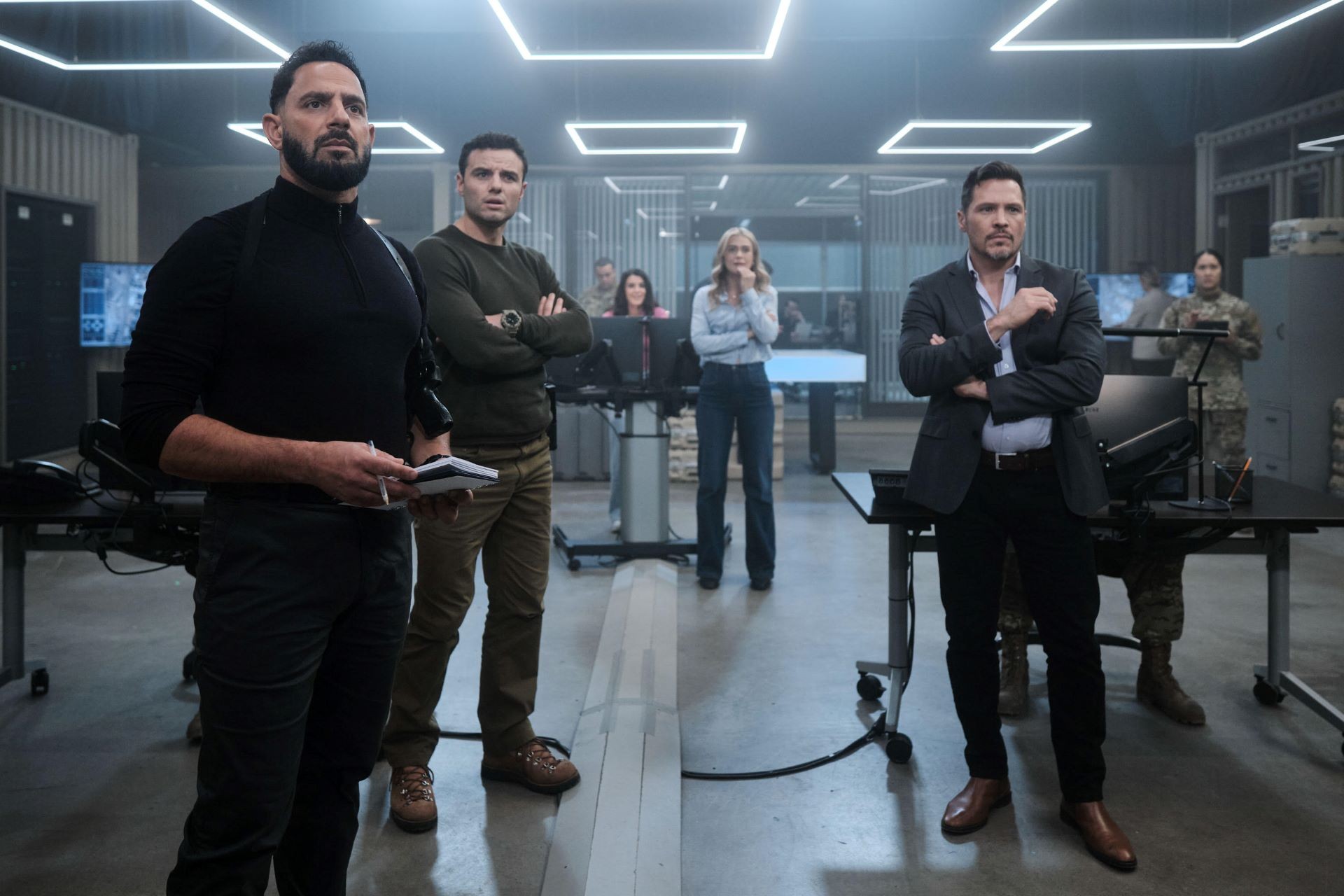 A group of people stand in a high-tech operations room, looking focused and serious.