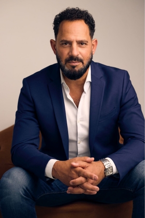 A man in a navy blazer and white shirt sits with his hands clasped, looking confidently at the camera.