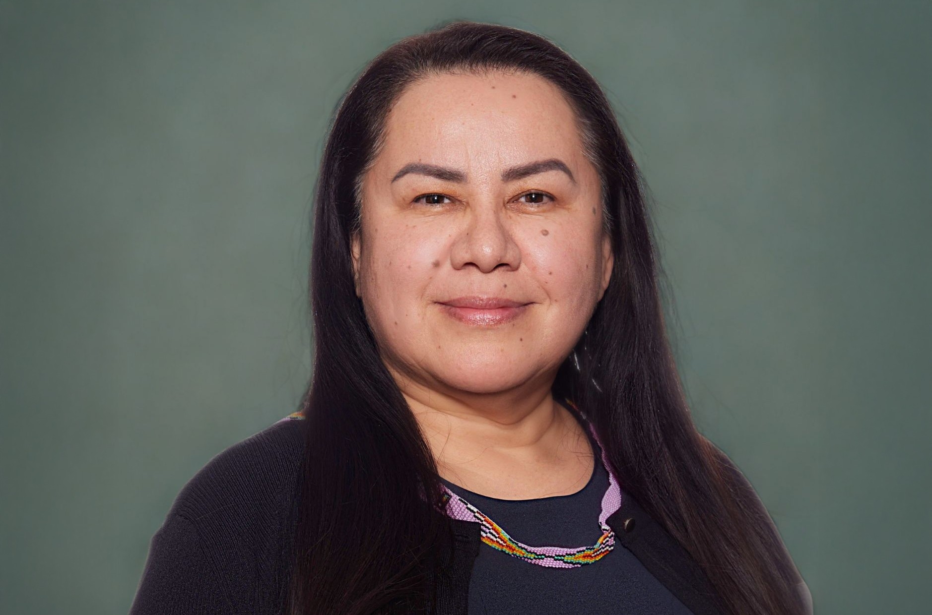 A woman with long, black hair is wearing a dark top with a beaded necklace.