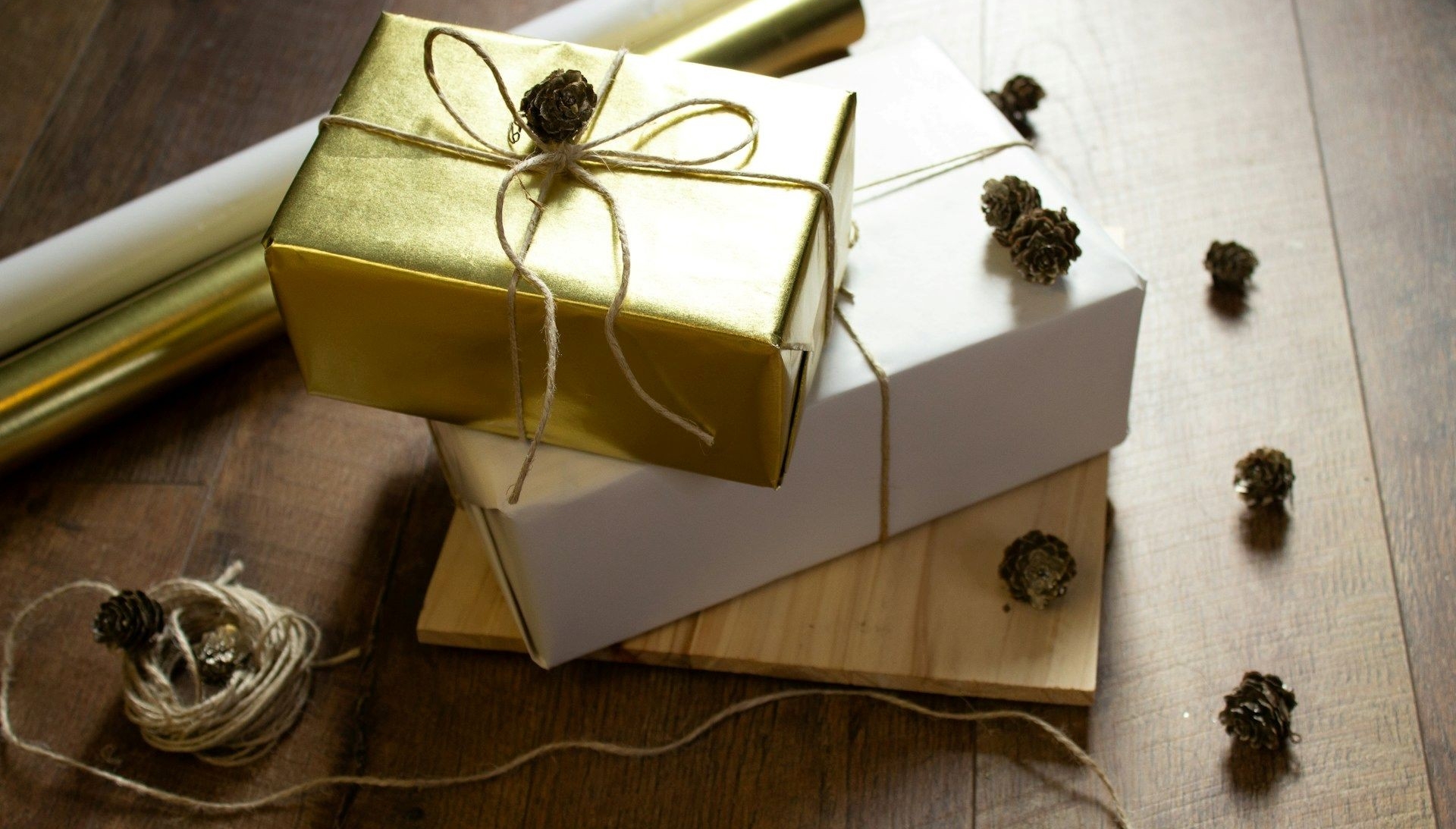 three gifts stacked up, one wrapped in gold paper, another in white paper, with jute string and pinecones lying on the table