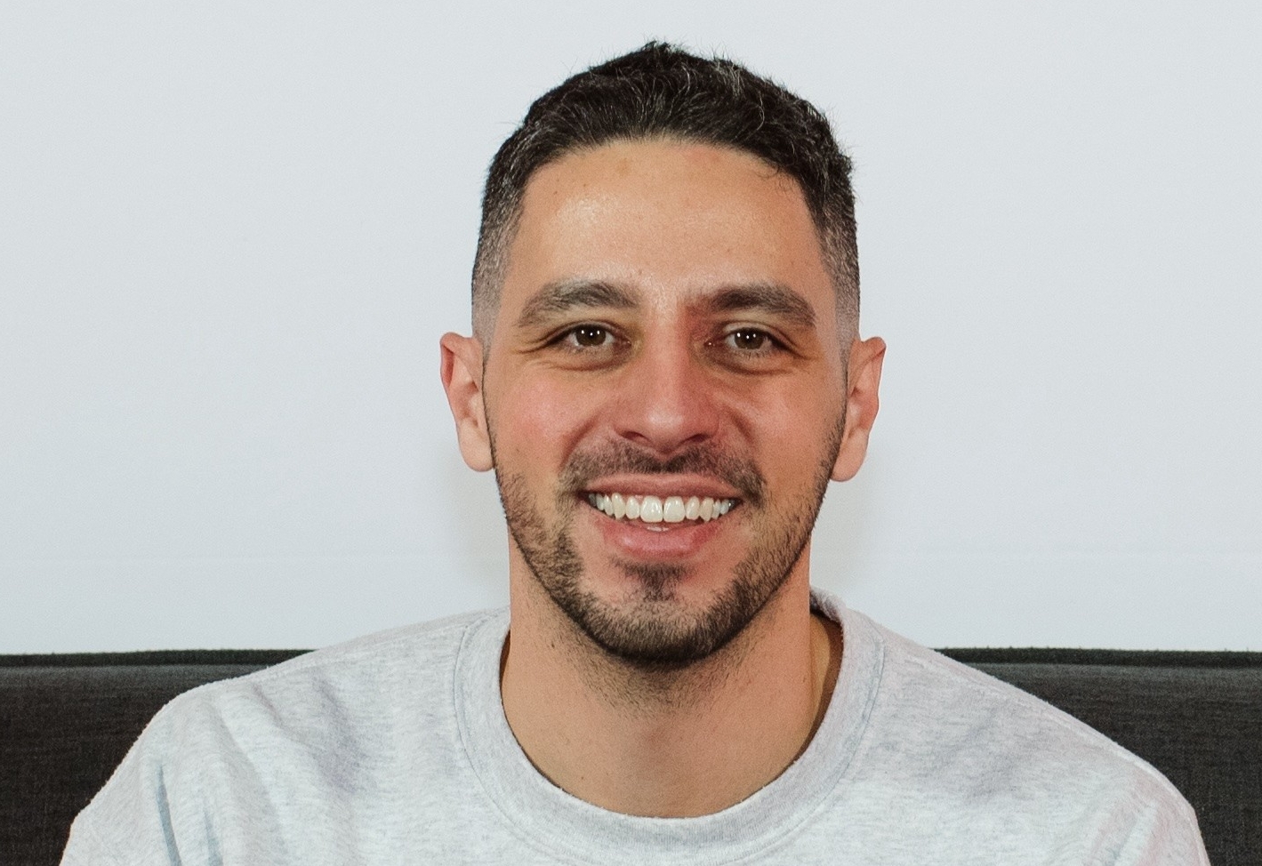 Man with short dark hair and a beard and wearing a gray T-shirt smiles and looks at the camera. 