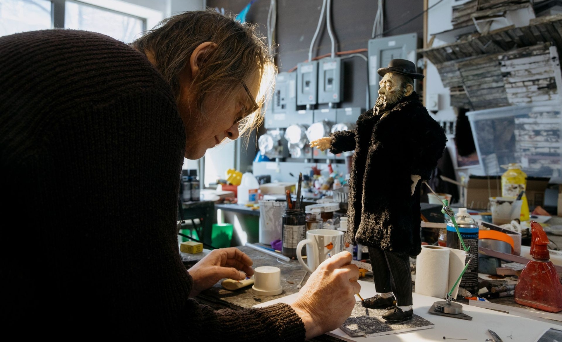 A man at a workbench in art studio leans over a pupper while painting detail on its feet. 