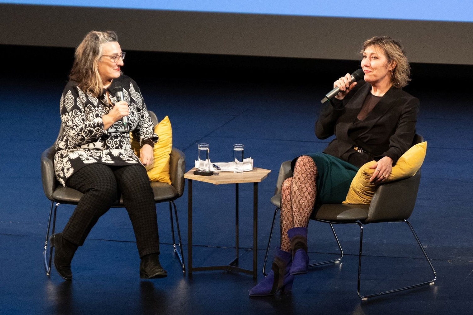 Cecil Castelluci and Martha Wainwright sit on chairs on stage. They hold microphones. There is a slideshow in the background.