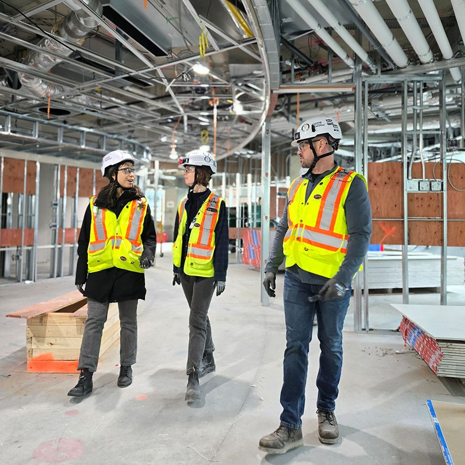 Two women and a man wear safety vests and hard hats as they walk through a contrustion site.