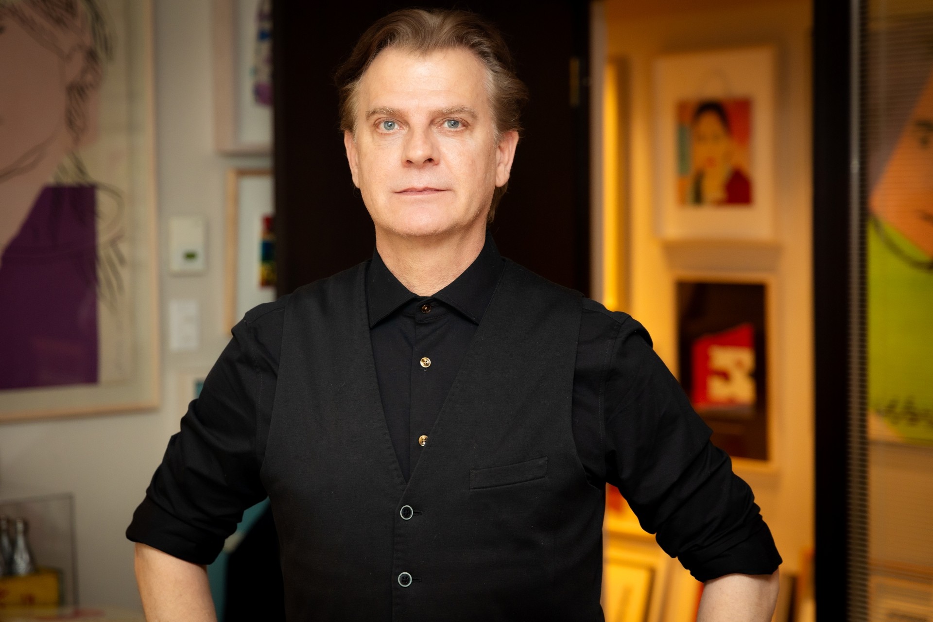 A man wears a black dress shirt and vest and stands in a room filled with art.