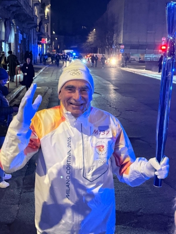 Man wearing an olympic tuque and track suit holding an Olympic torch runs down a city street in the evening