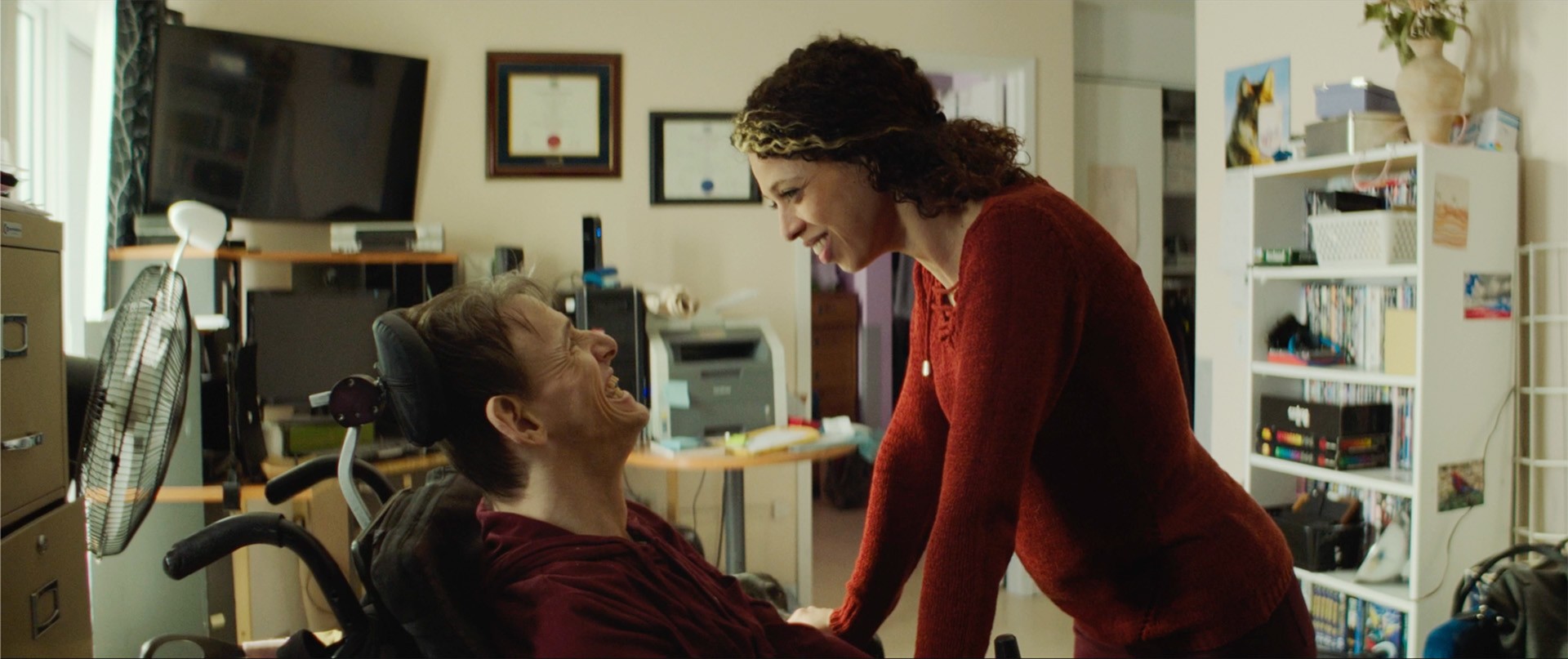 A smiling woman leans over a laughing man who is wheelchair-bound.