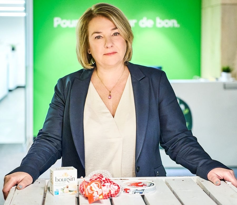 Cristine stands behind a table uon wihich are displayed cheese products: Boursin, Babybel and Laughing Cow.