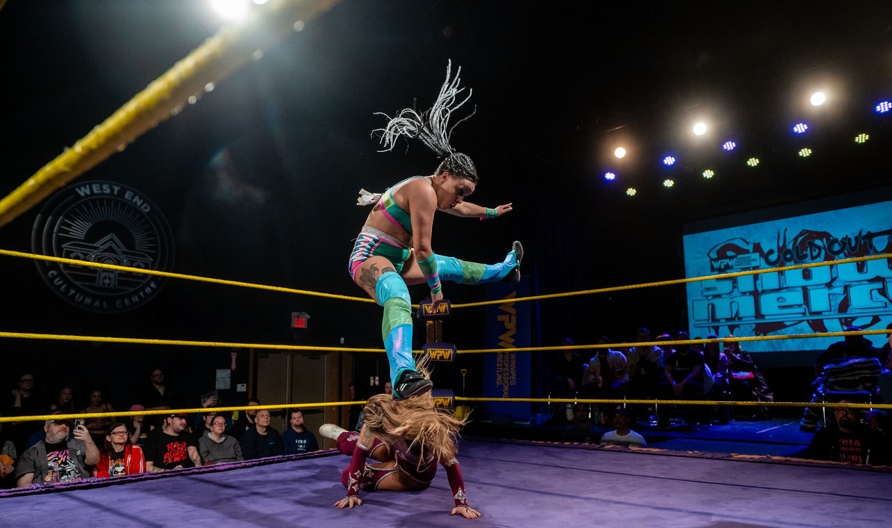 Wrestler Kristara jumps over her opponent, who has fallen to the ground, in the ring,