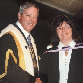A man and a woman dressed in ceremonial graduation robes shake hands and smile.