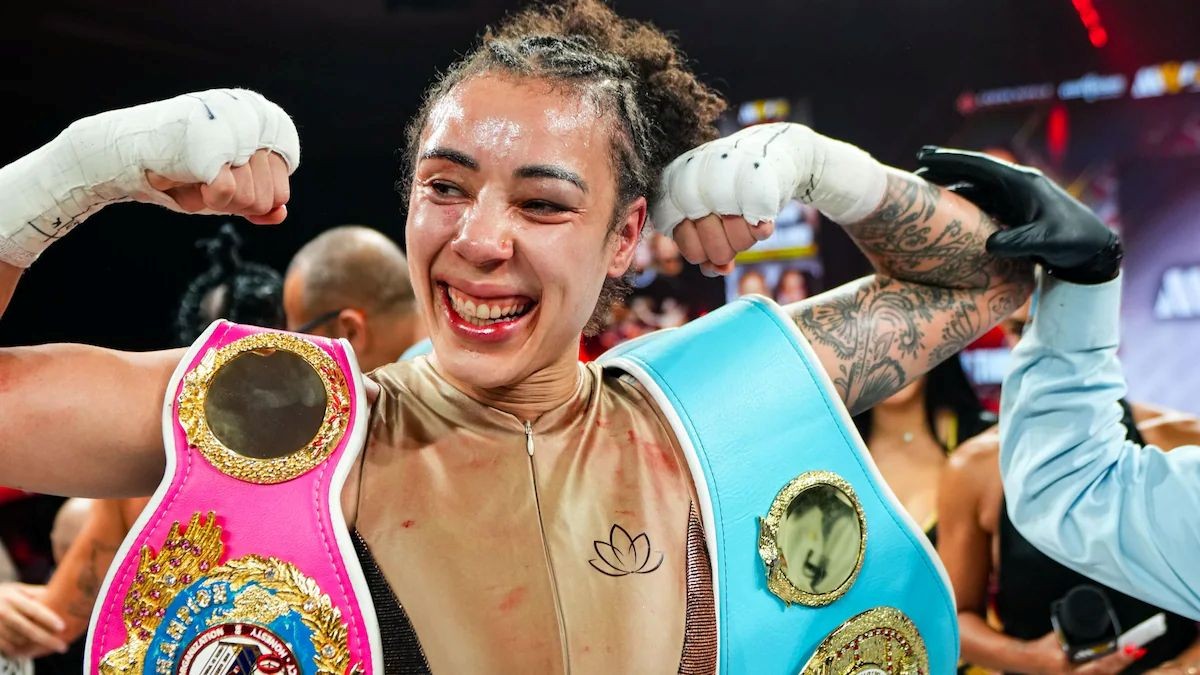 After her champion fight, smiling Tammara holds her arms up in victory while a championship belt is draped over each shoulder.