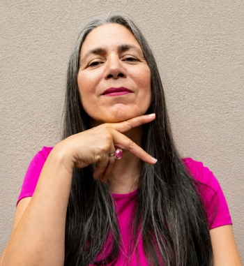 Skawennati makes a "peace sign" with her right hand under her chin. She is wearing a pink t-shirt and has long hair.