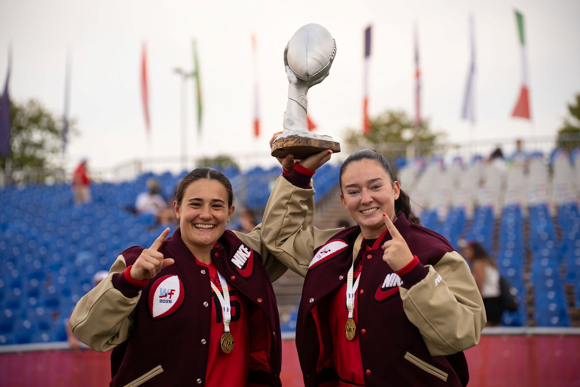 Sara and Caroline are each wearing varsity jackets and winners medals while holding a trophy above their head.