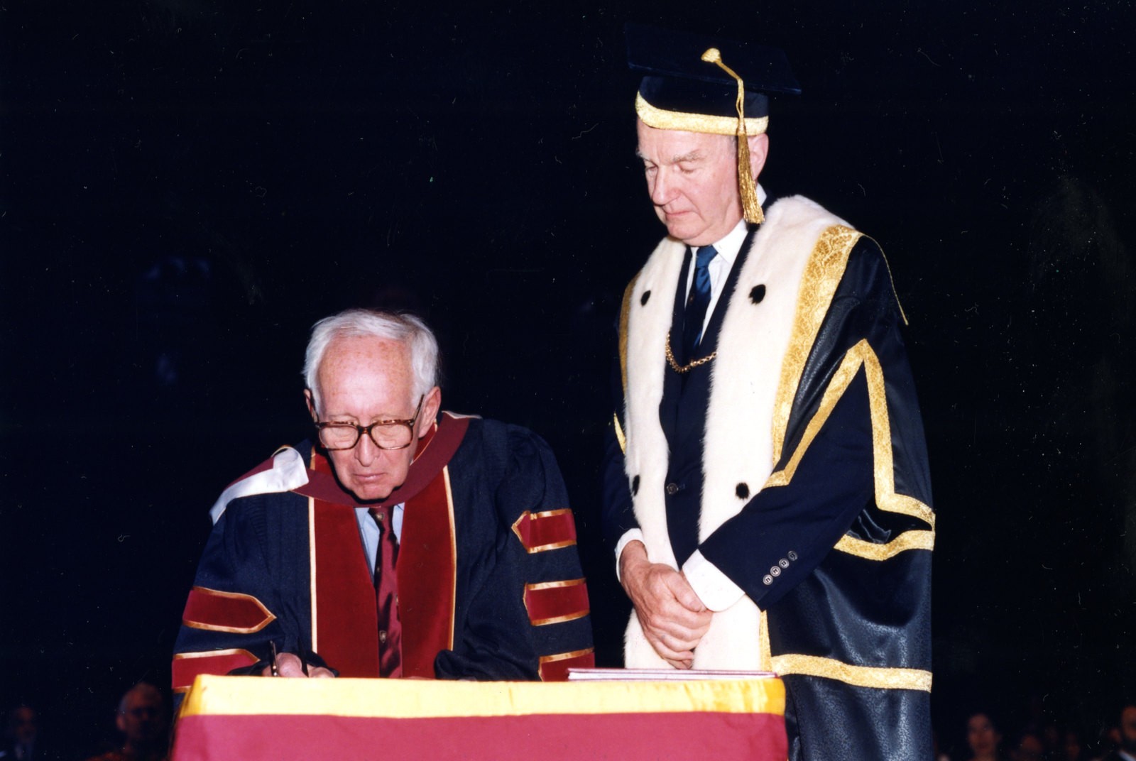 Two men in conovcation robes are side-by-side, one is signing a book as the other looks on.