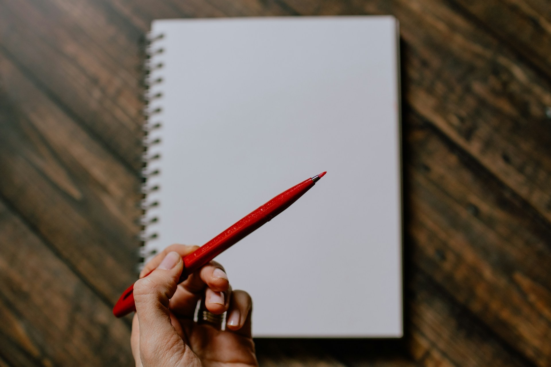 A bank white page in a spiral notebook with a hand above it holding a red pen