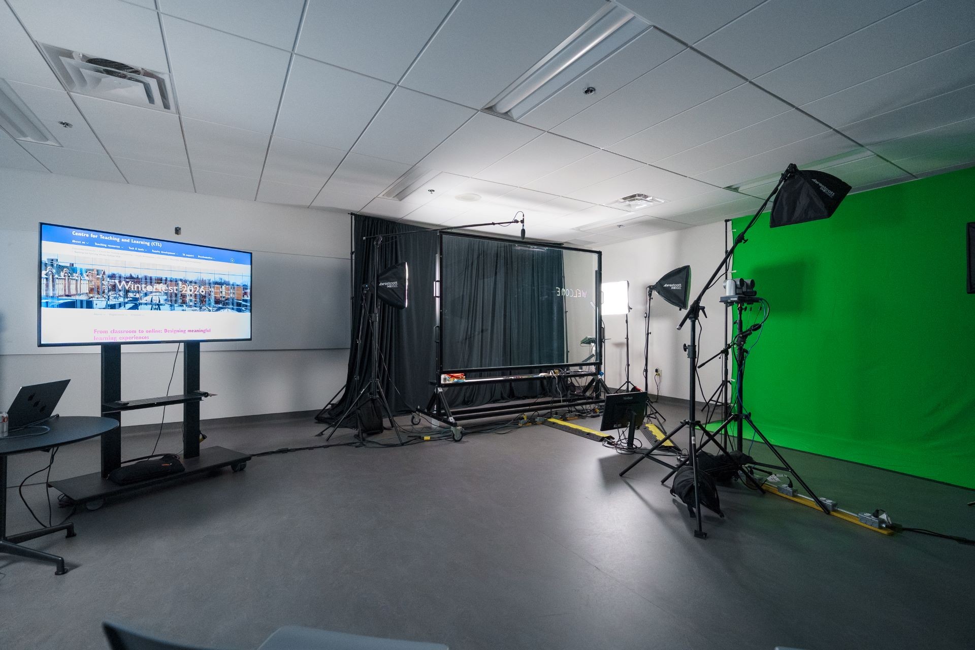 Photo of the lightboard studio space showing a large clear glass writable standing screen on the right side of the photo surrounding by large light stands and a green green and a TV screen showing the CTL Winterfest 2026 website in a large monitor on the left side of the photo.