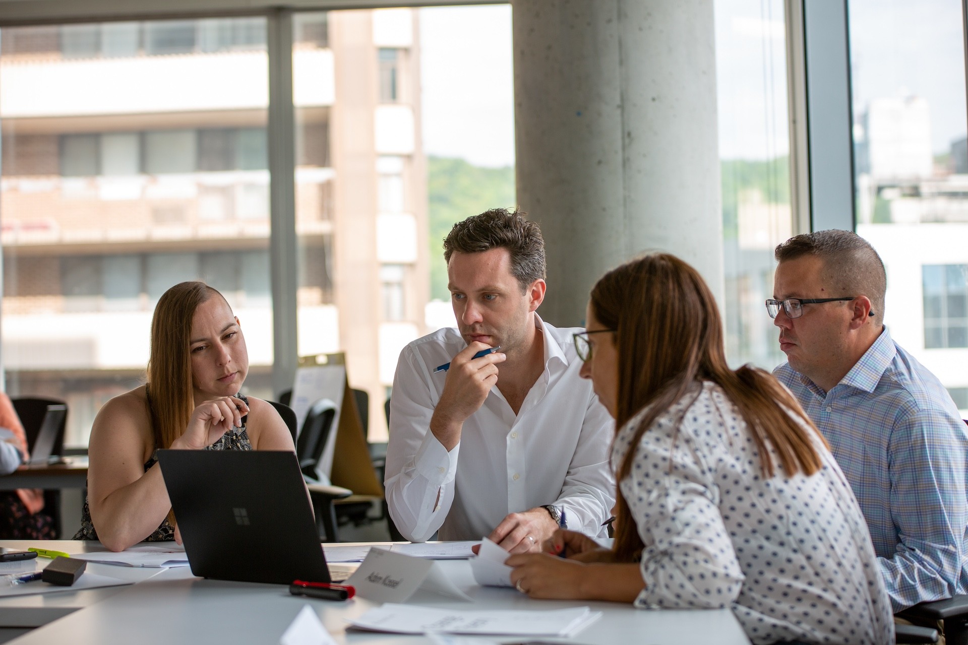 Four people seated at desks looking at documents on the desk with windows in the background with outside view of buildings and sky
