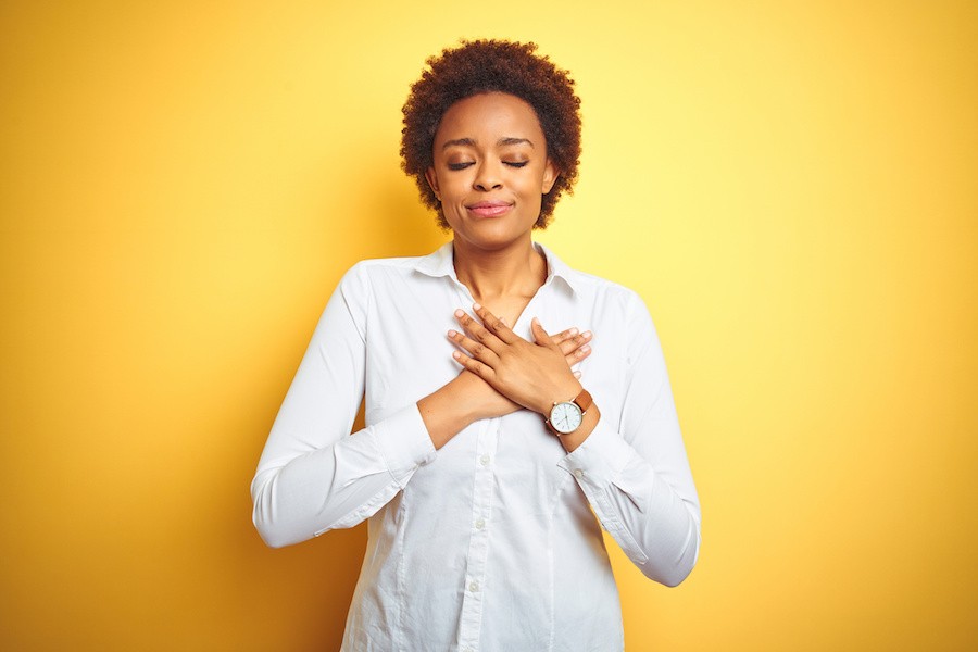A woman with a plain yellow background putting her hands on her heart