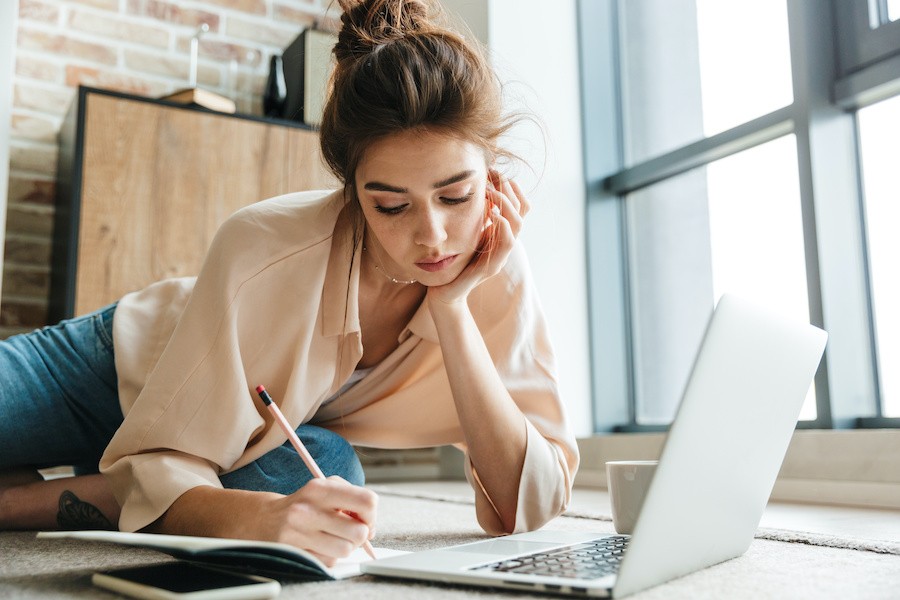 a woman writing in a notebook in front of a laptop with hr hand under her chin and thinking