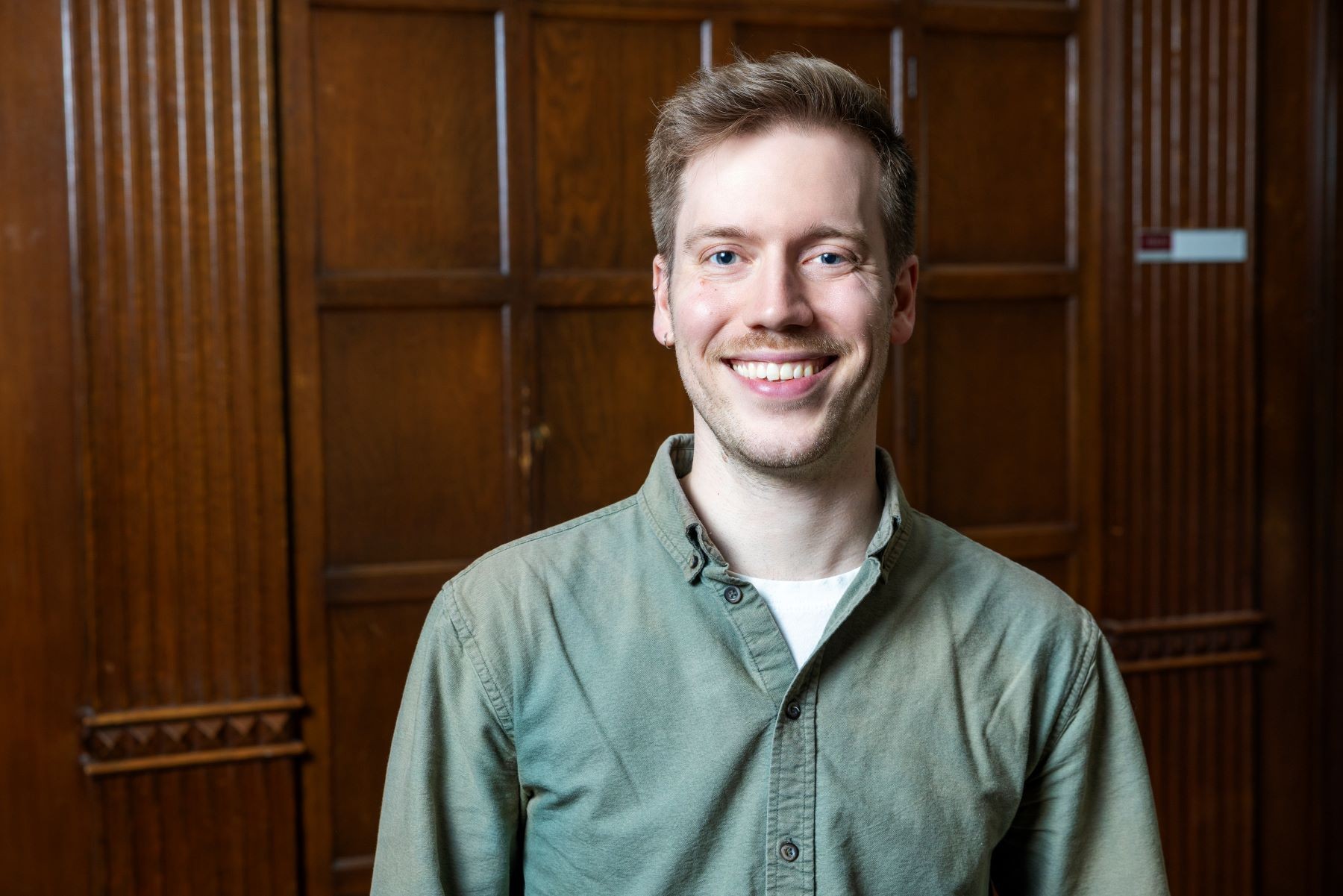 Nick Grunden A young man with blonde hair and pale skin, and wearing a sage green button-up shirt, smiles in front of a wooden wall.