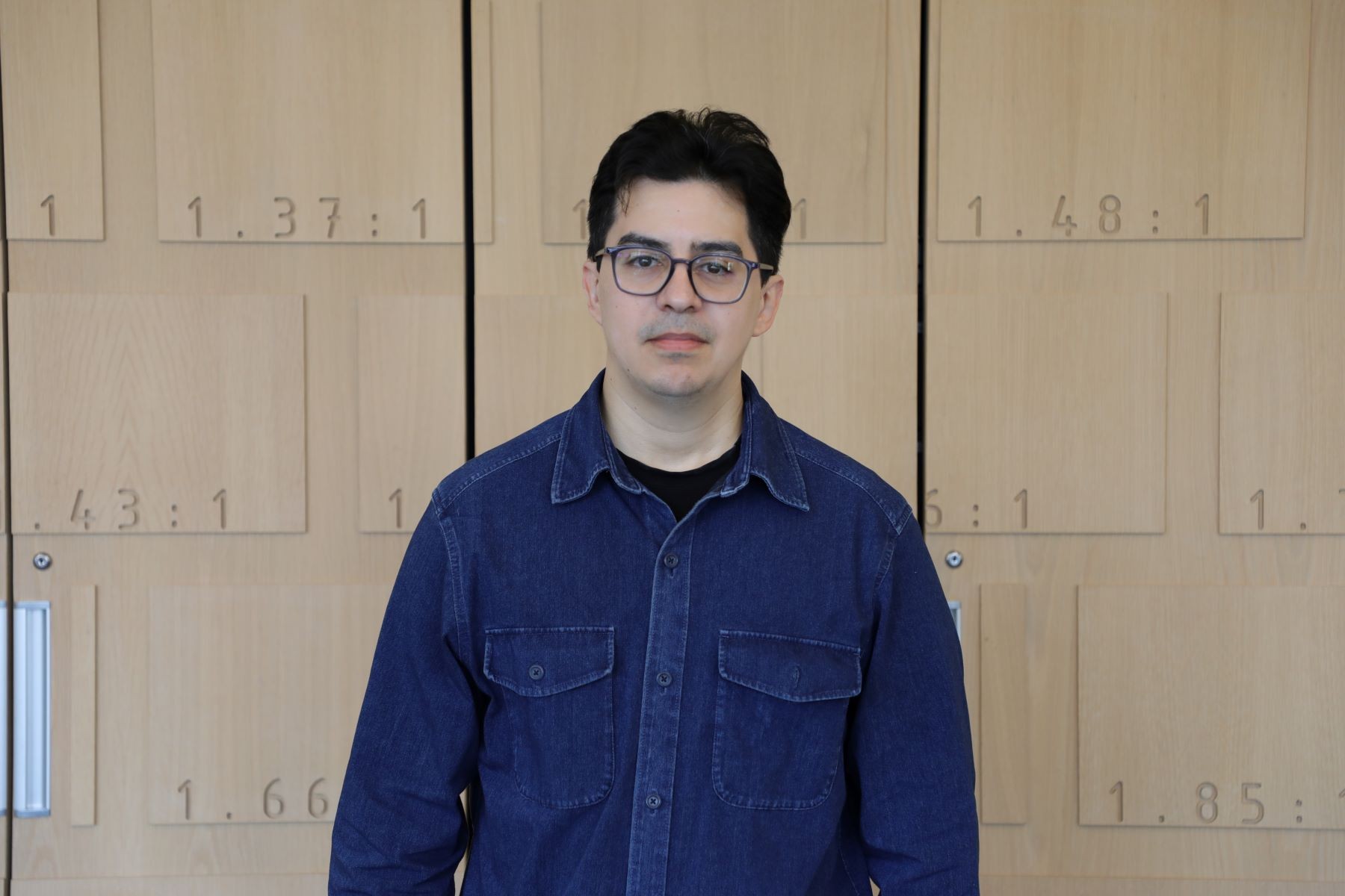 Vinicius Sanchez A man with black hair and pale skin wearing dark-rimmed glasses and a jean button-up shirt poses for the canera.