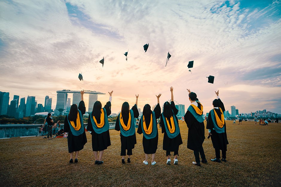 Seven students wearing black graduation robes and orange and blue sashes throwing mortarboards into the air. They are facing the other way looking at the city while standing in a green field.