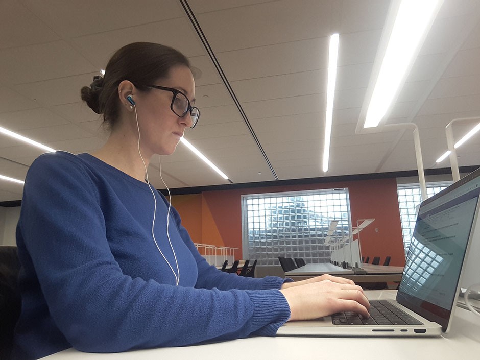 Andrea Cartile is pictured here from the right side working in the Concordia University Webster Library in downtown Montréal. She has brown hair tied in a bun, is wearing glasses and a dark blue long-sleeved shirt. She is seated, facing a laptop, and wearing wired headphones.