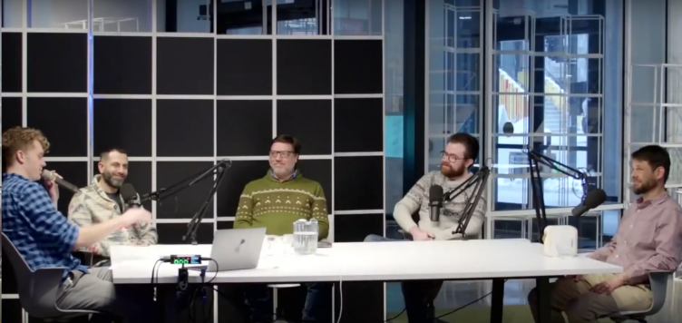 A group of panelists sit around a table, each holding a microphone.