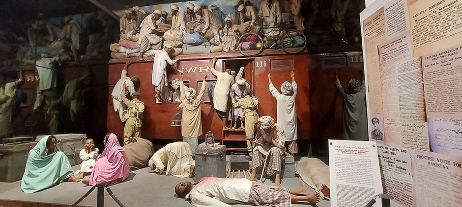 A group of people, mostly men, wearing traditional dhoti and kameez standing on the roof of a train and on the platform unloading their luggage. Pakistani army officers can be seen helping them get off the train. One man can be seen injured lying, while two women and a child also sit on the platform.